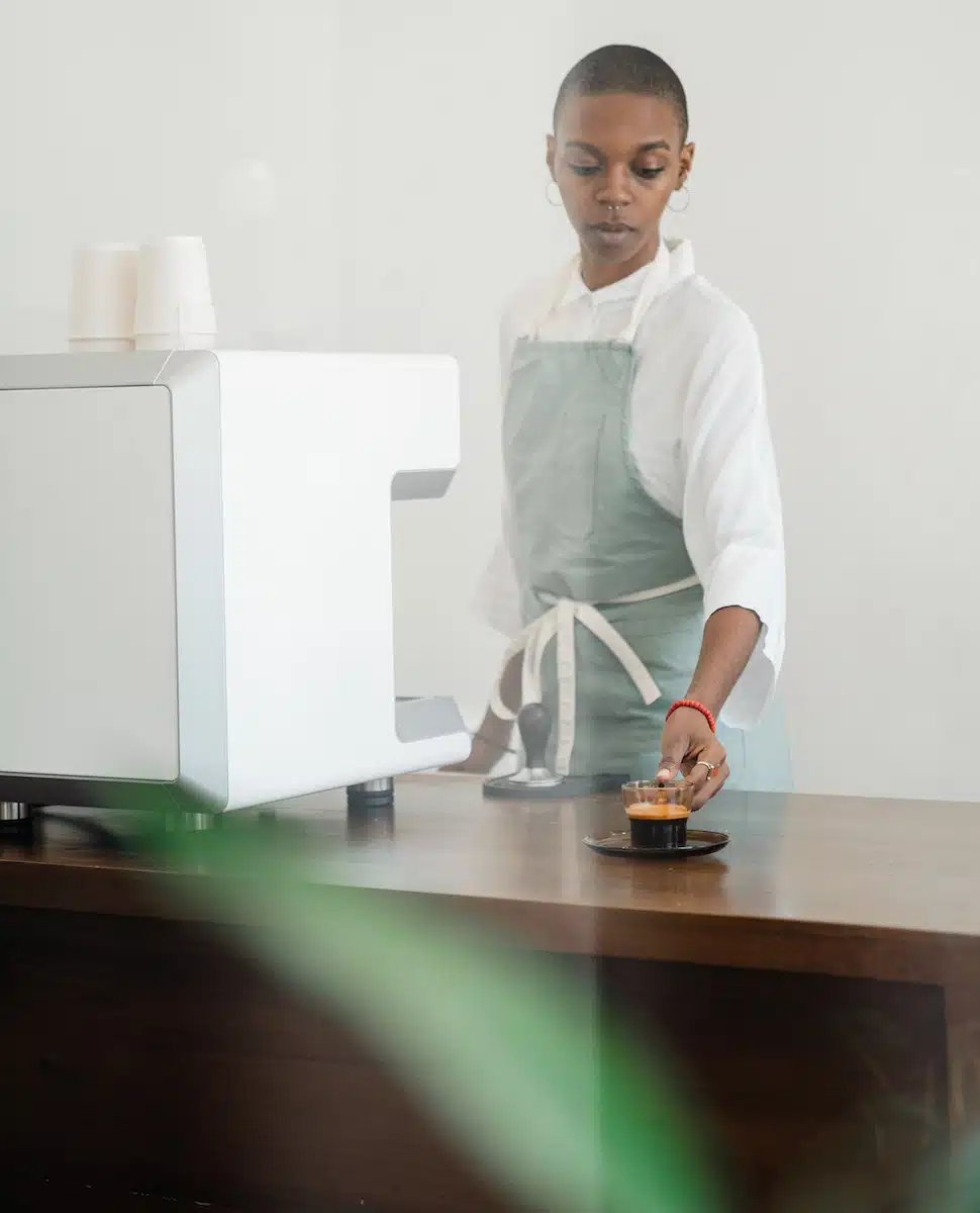 Concentrated African American female barista in apron standing nearby professional coffee machine and serving hot aromatic espresso in transparent cup in light cafe