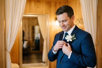 Marié en costume bleu marine attachant une boutonnière fleurie sur son revers devant un miroir lors de la préparation du mariage