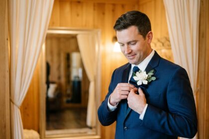 Marié en costume bleu marine attachant une boutonnière fleurie sur son revers devant un miroir lors de la préparation du mariage